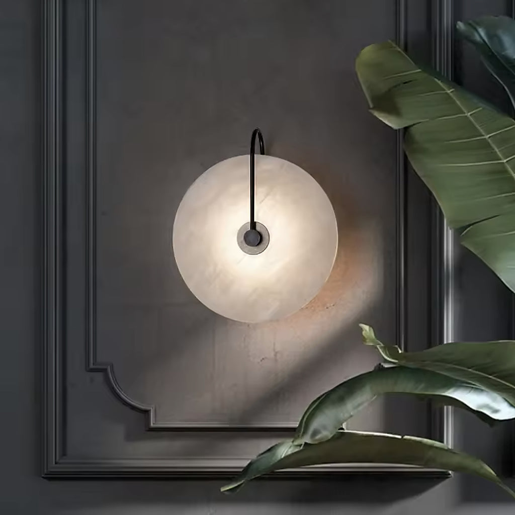 Alabaster disc wall sconce on a dark panelled wall beside foliage